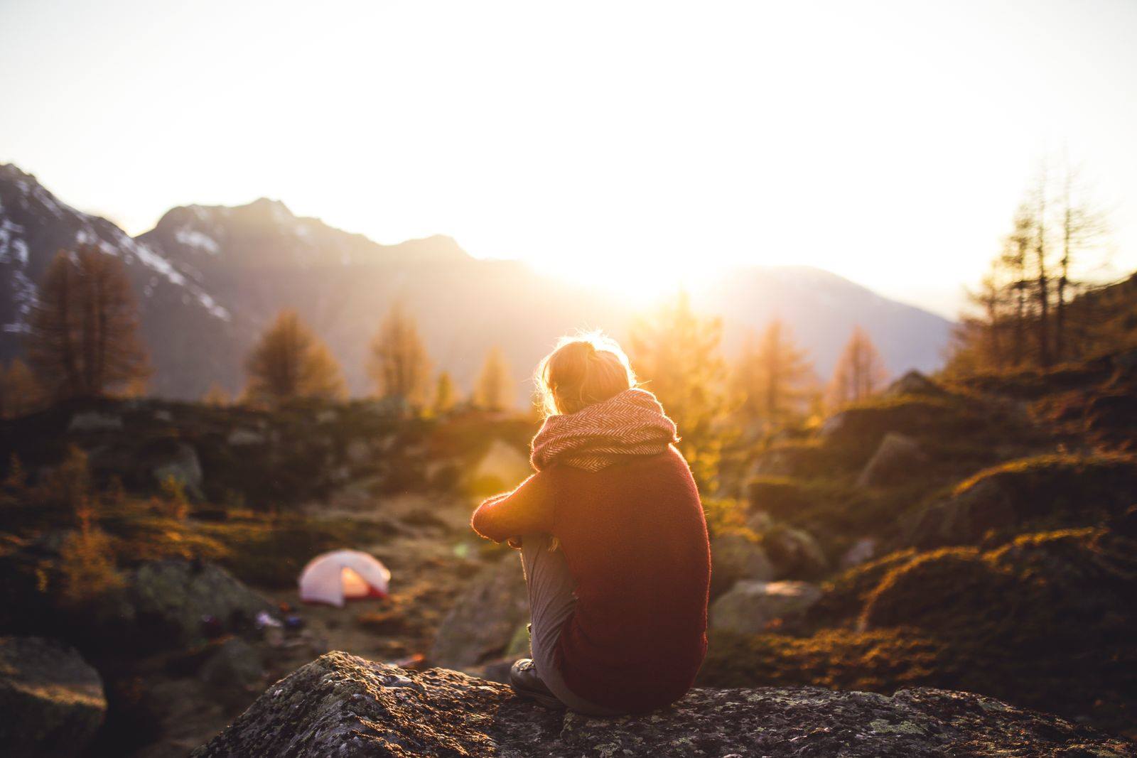 A woman sitting alone on a rock, looking out at a mountain view at sunrise
